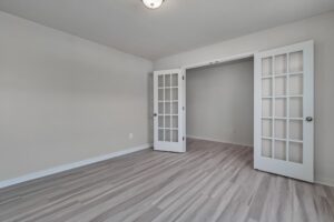 An empty room with light gray wooden floors, white walls, and double French doors with glass panels. The ceiling has a central light fixture.