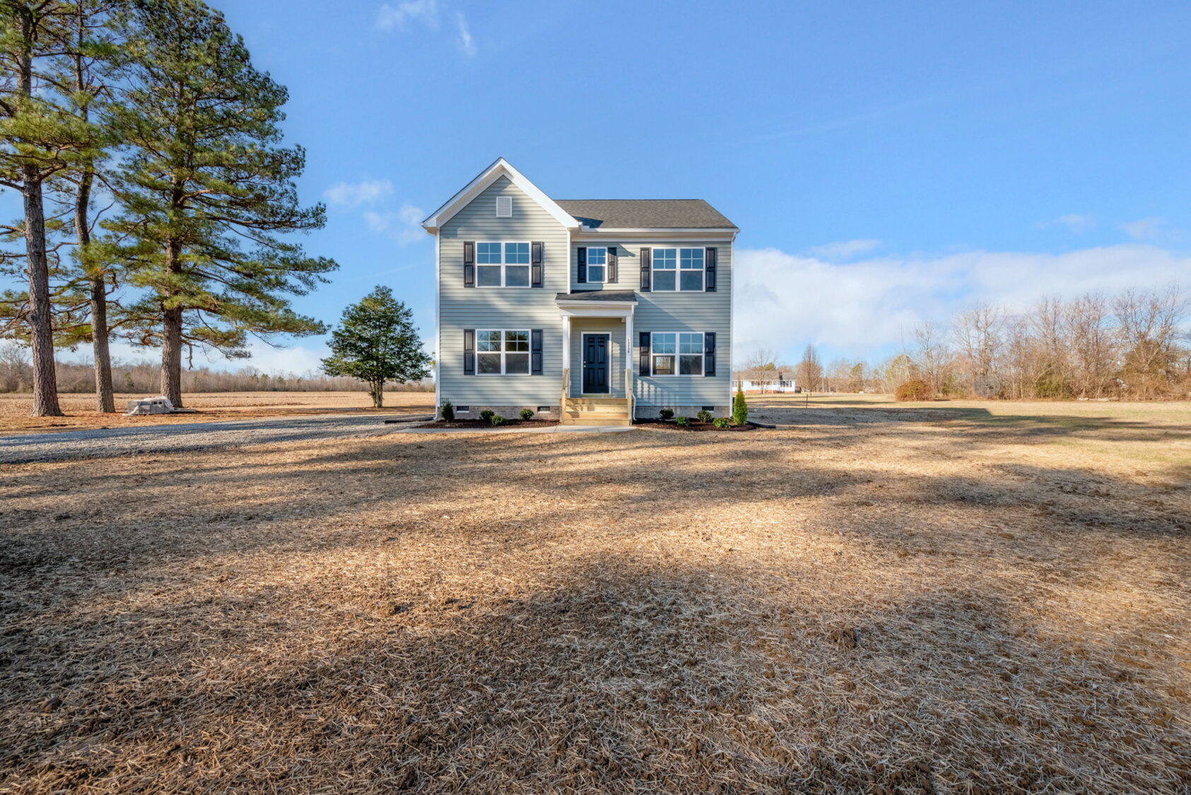 White two-story house with blue shutters on a large, open lot, surrounded by trees under a blue sky.