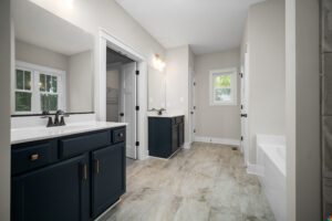 A modern bathroom with dual sinks, navy cabinetry, a large mirror, beige tiled floor, a window, and a bathtub.