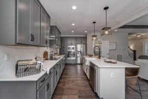Modern kitchen with grey cabinets, white countertops, and stainless steel appliances. An island with chairs, pendant lights, and wood flooring complete the space.