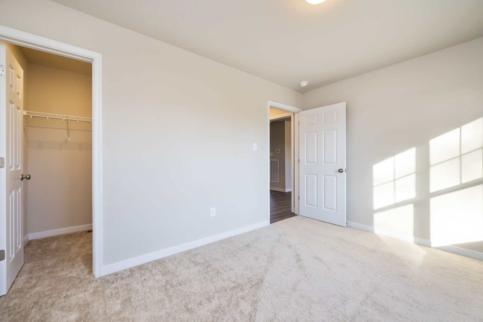 Bright, empty bedroom with beige carpet, white walls, closet, and sunlight streaming through a window.