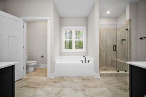 A modern bathroom with a bathtub, shower with glass door, toilet, and double sink vanity. The room has beige tiles and a window above the bathtub.