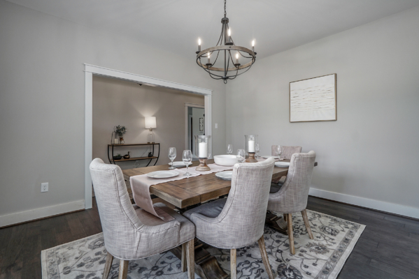 A dining room with a wooden table set for six, upholstered chairs, and a chandelier. The walls are light gray, and there's a framed art piece. An adjacent room is visible through an open doorway.