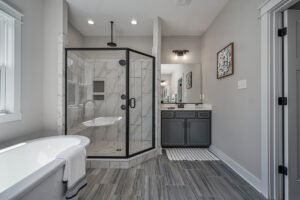 Modern bathroom featuring a glass-enclosed shower, a white freestanding bathtub with a towel, dual-sink vanity with mirror, grey cabinets, and wall art. The floor has grey tiles.