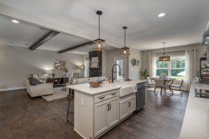 Open-concept kitchen and living room with exposed beams, modern light fixtures, a central island with a farmhouse sink, and a dining area with large windows letting in natural light.