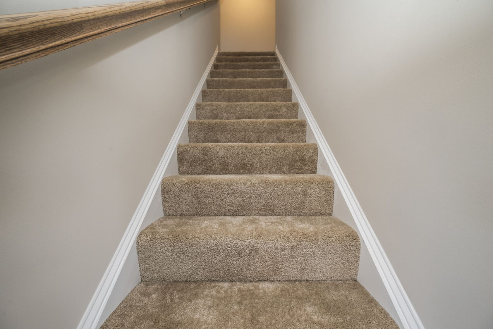 Carpeted staircase with beige walls and a wooden handrail, leading up to a well-lit hallway.