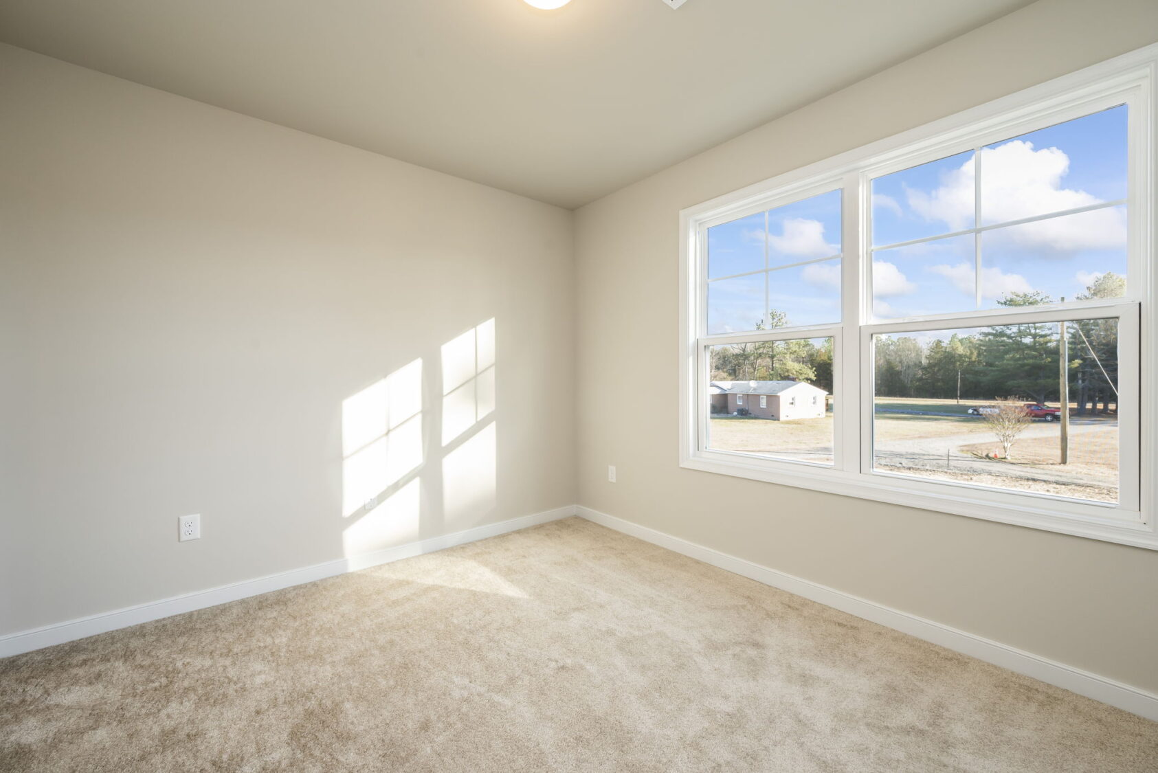 Empty room with beige carpet, light-colored walls, and a large window letting in natural sunlight.