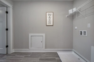 A laundry room with tiled flooring, a white shelf, hookups for appliances, and a framed sign that reads "WASH" on the wall.