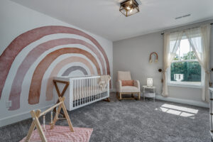 A nursery with a rainbow mural, white crib, rocking chair, small side table with a lamp, and a play gym on a pink rug. Sunlight streams in through a window with sheer curtains.