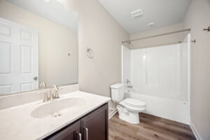 A modern bathroom with white fixtures, including a bathtub with a shower, a toilet, and a sink with a large mirror above it, featuring wooden flooring.