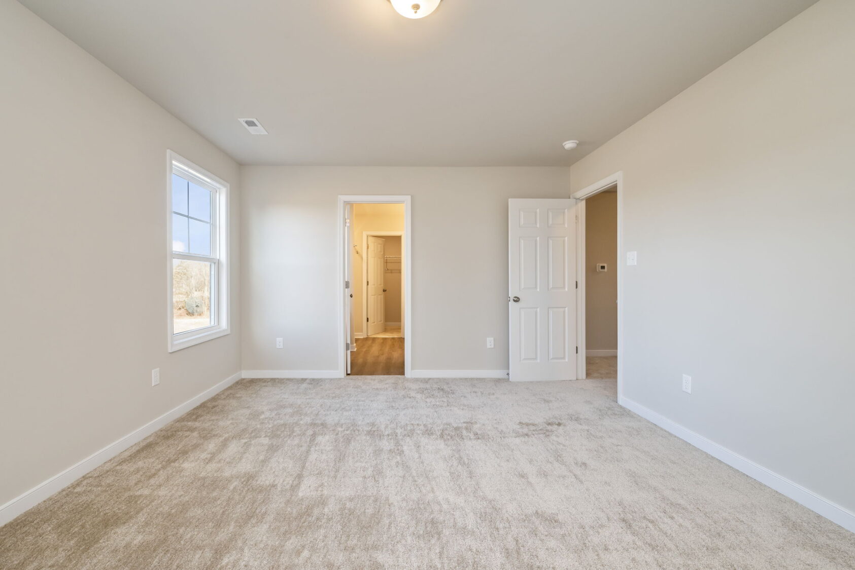 Empty beige carpeted bedroom with two doorways, one open door, and a window letting in natural light.