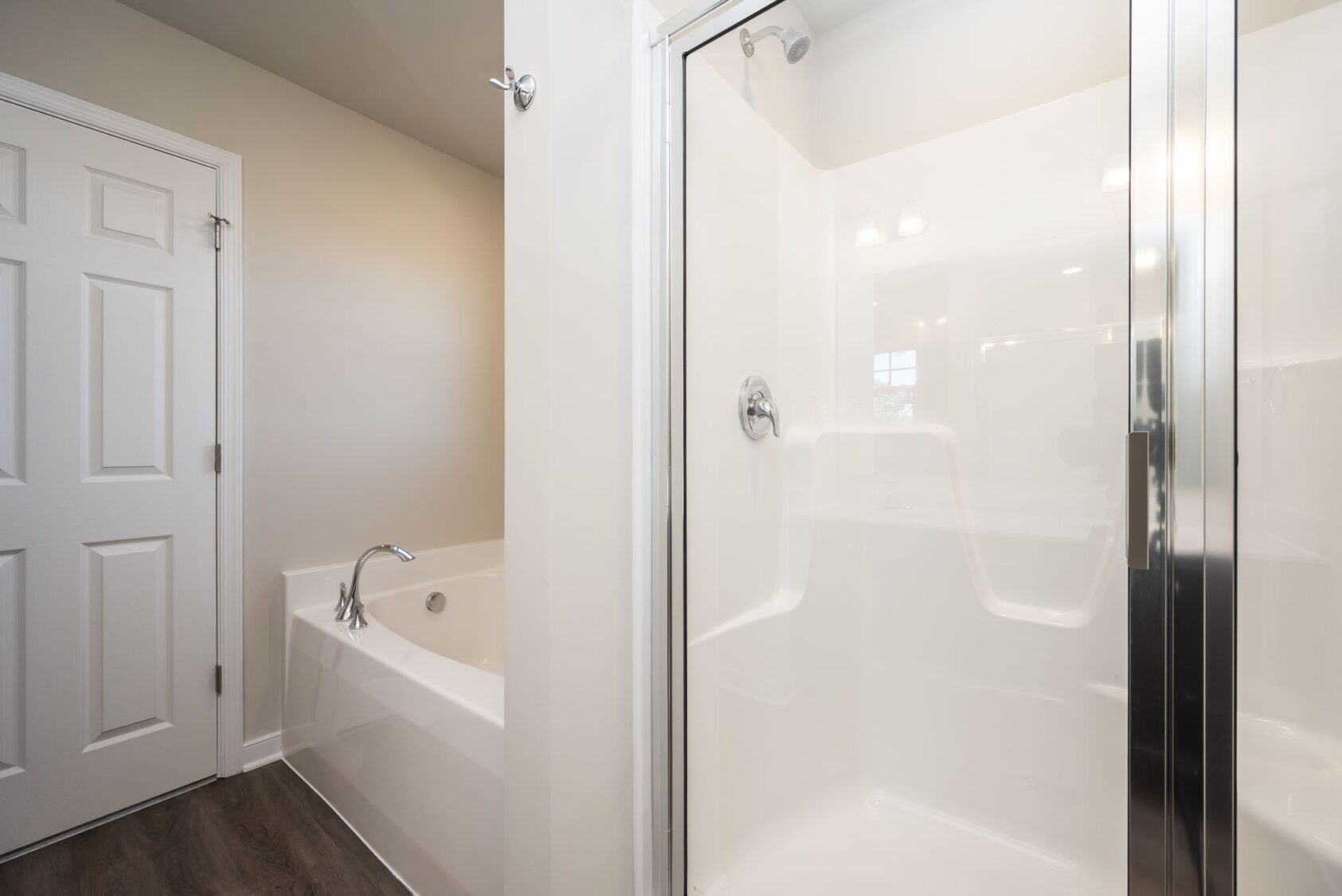 Modern bathroom with a glass shower, white bathtub, wood floor, and beige walls.