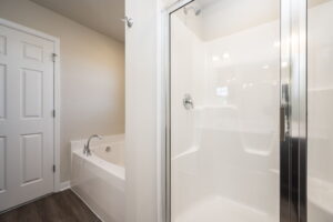 Modern bathroom with a glass shower, white bathtub, wood floor, and beige walls.
