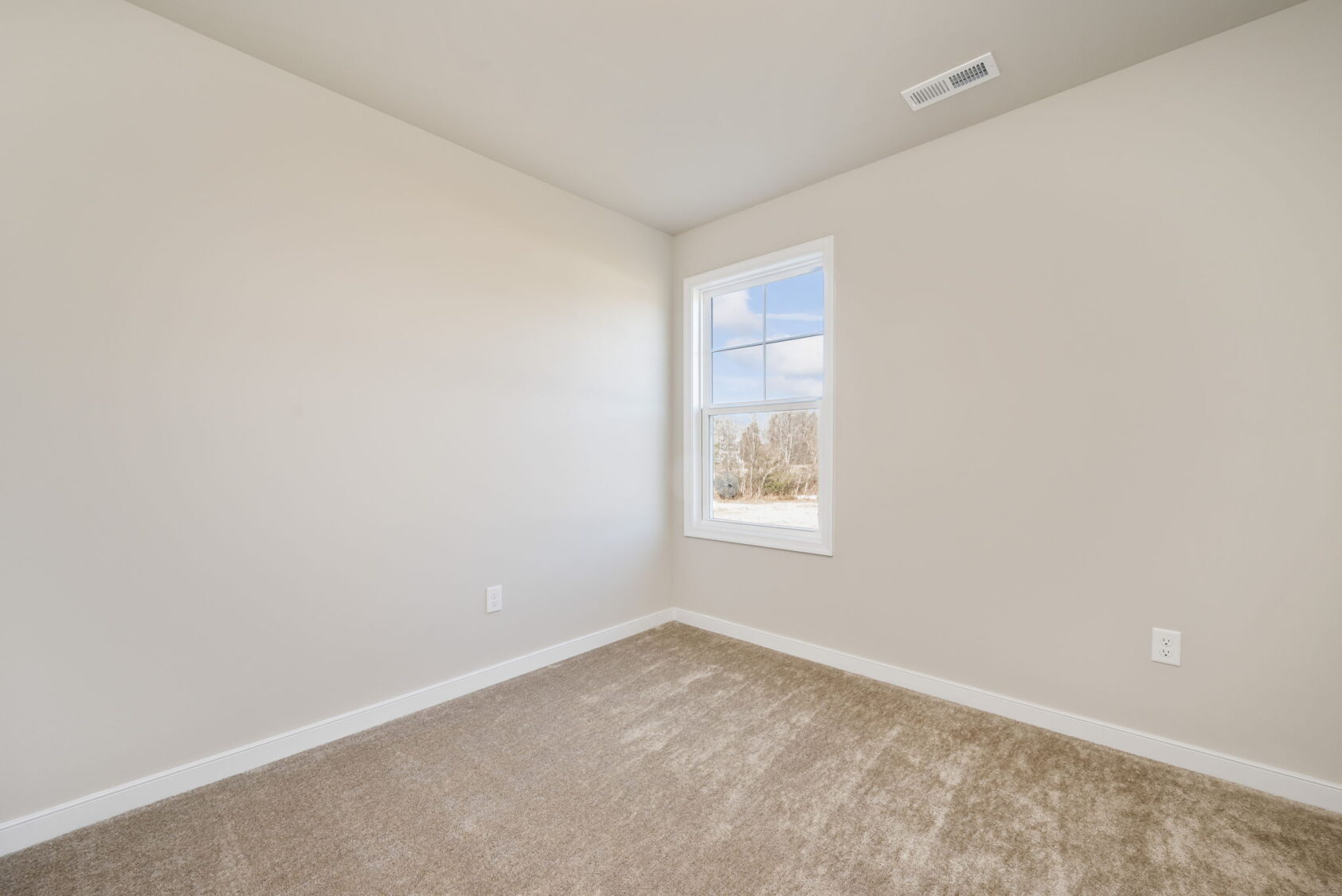 Empty beige room with carpet flooring, a single window, and white walls with electrical outlets.
