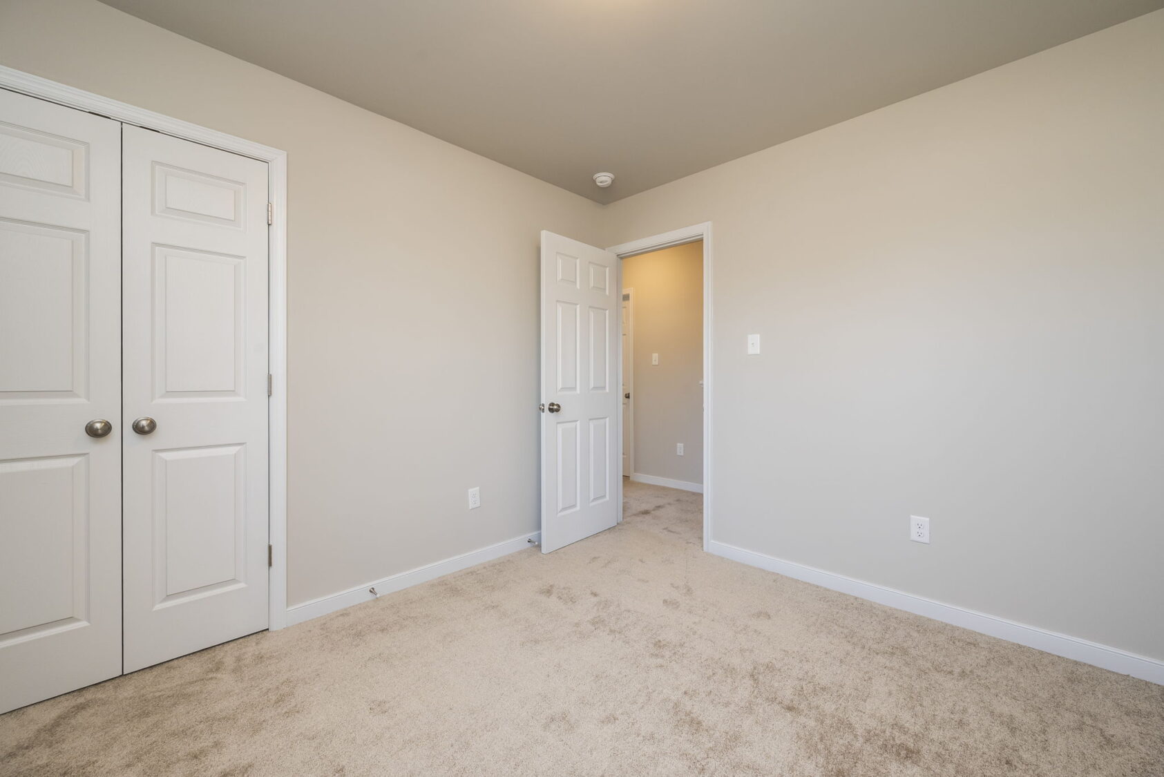 Empty beige room with carpet, double closet doors, and an open door leading to a hallway.