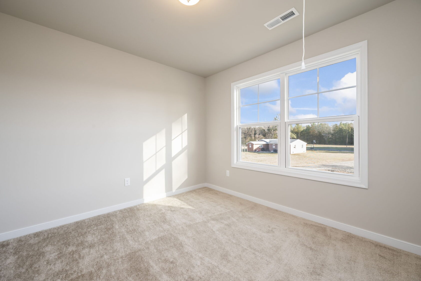 Empty, sunlit room with beige carpet, light walls, and a large window showing a clear view outside.