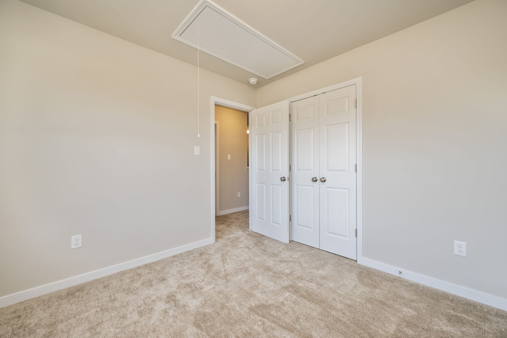 Empty beige carpeted room with double white doors, light walls, and an attic access panel in the ceiling.