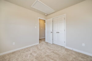 Empty beige carpeted room with double white doors, light walls, and an attic access panel in the ceiling.