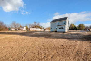 A gray two-story house sits on a large, bare dirt lot under a clear blue sky with scattered clouds.