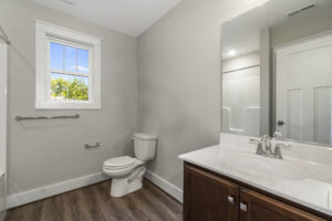 A bathroom with a white toilet, a wooden vanity with a white countertop and sink, a large mirror, and a window. The floor has wooden flooring, and the walls are painted light gray.