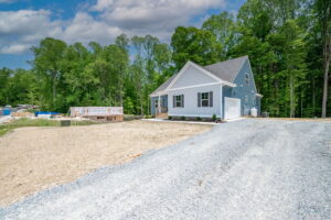 A new single-story house with white siding and gray shingles, featuring a gravel driveway and surrounded by a wooded area under a clear blue sky.