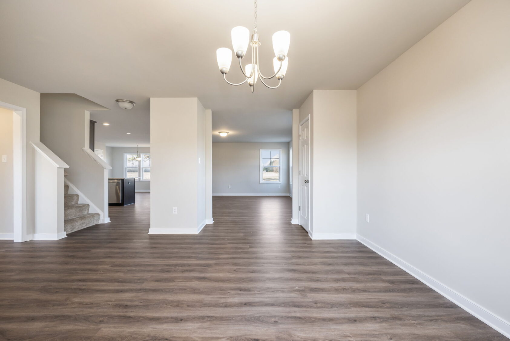 Bright, empty room with wood flooring, neutral walls, chandelier, and view of kitchen and stairs.