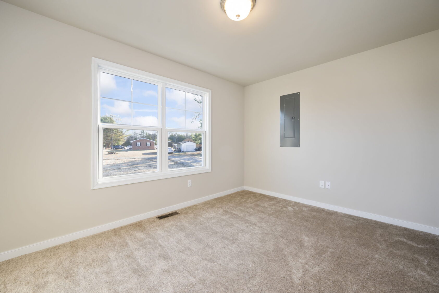 Empty carpeted room with a large window, beige walls, and an electrical panel on the right wall.