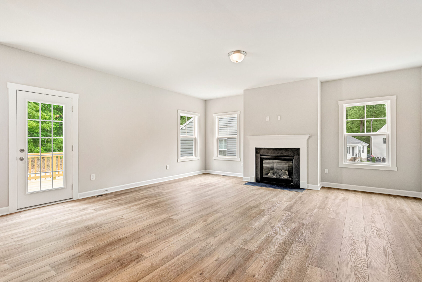 Bright, empty living room with wood floors, a fireplace, and windows overlooking a deck and green yard.