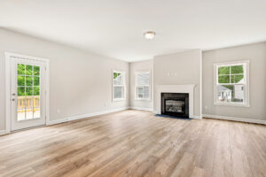 Bright, empty living room with wood floors, a fireplace, and windows overlooking a deck and green yard.