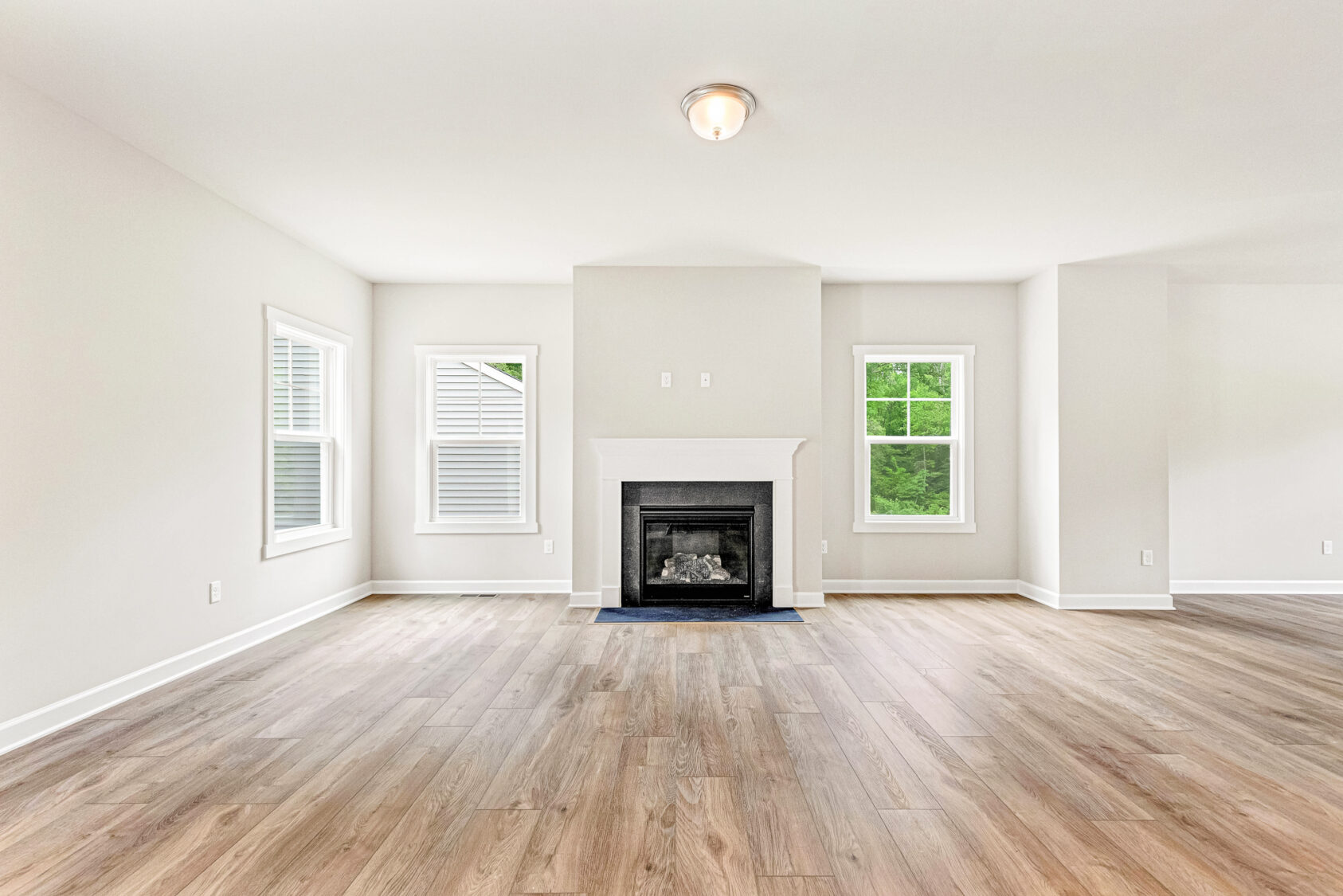 Bright empty living room with light wood floors, white walls, windows, and a central fireplace.