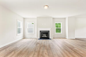 Bright empty living room with light wood floors, white walls, windows, and a central fireplace.