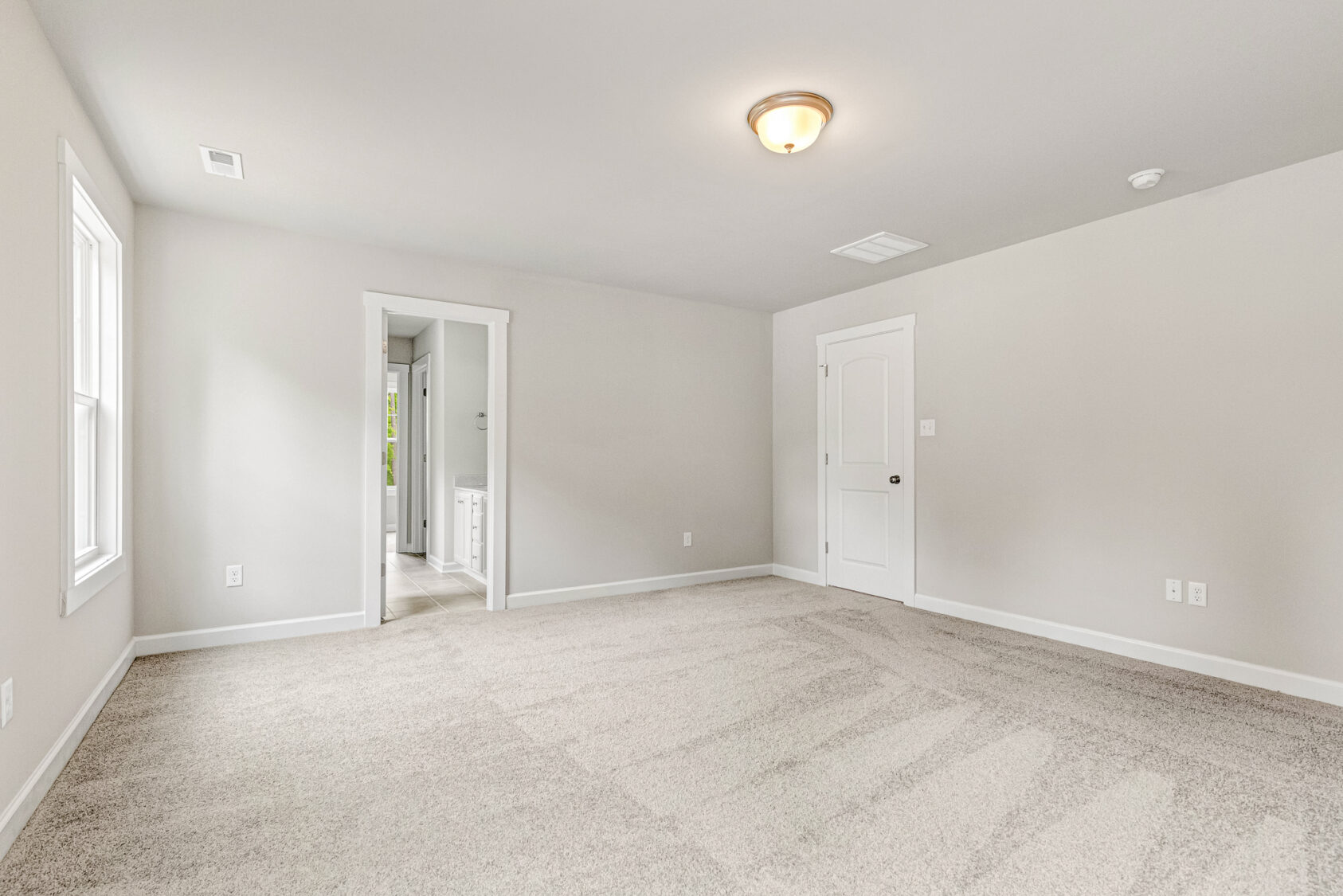 Empty carpeted room with beige walls, ceiling light, two doors, and a window letting in natural light.