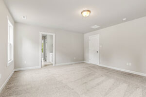 Empty carpeted room with beige walls, ceiling light, two doors, and a window letting in natural light.