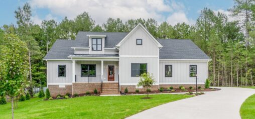 A white, modern farmhouse-style house with a gray roof and front porch, surrounded by a green lawn and trees, is shown on a sunny day. A curved driveway leads to the entrance.