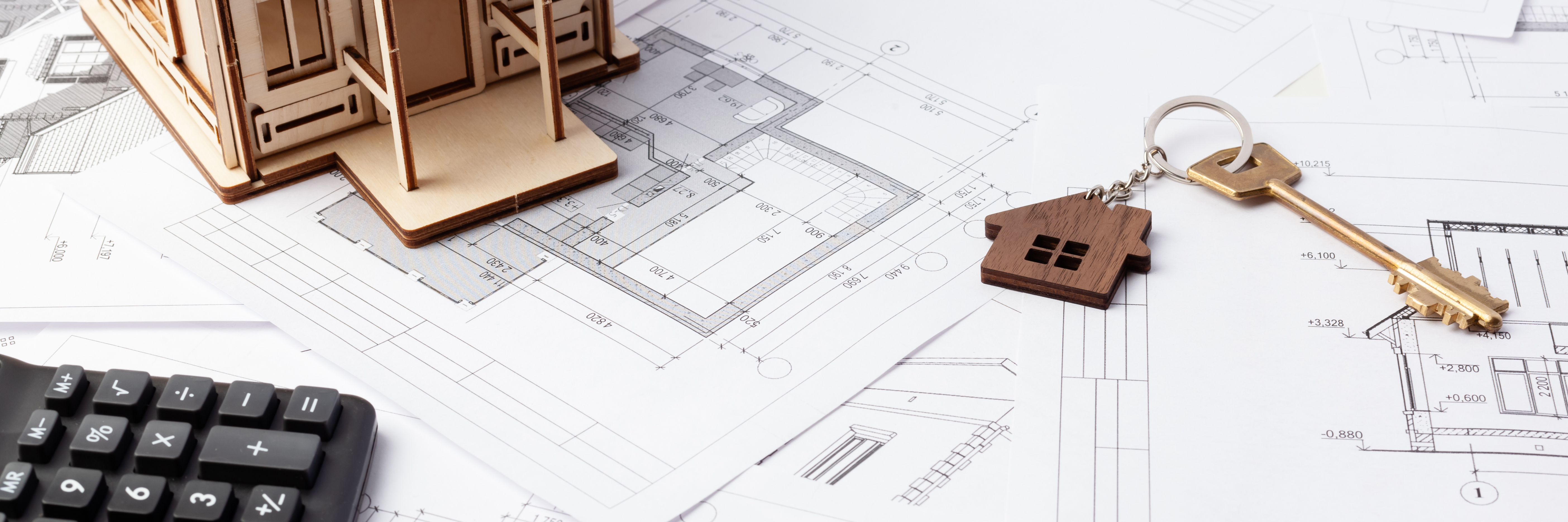 A wooden house model, architectural blueprints, a wooden house-shaped keychain with a key, and a calculator are spread out on a table.