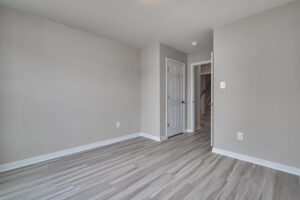 A small empty room with light gray walls, light wood laminate flooring, a white baseboard, a closed white door, and an open doorway leading to a staircase.