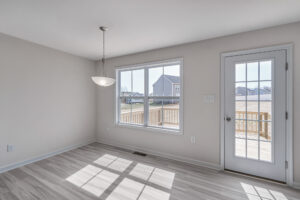 A bright, empty room with light-colored flooring, a hanging light fixture, a large window, and a glass door leading to a wooden deck outside.