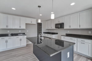 A modern kitchen with white cabinets, black countertops, a center island with a sink, pendant lighting, and a built-in microwave. The kitchen features light wood flooring and recessed ceiling lights.