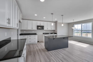 A modern kitchen with white cabinets, black countertops, an island with a sink, pendant lights, a microwave, and a large window providing natural light.