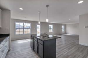 Modern open-concept kitchen and living area with an island, stainless steel sink, pendant lighting, and large windows providing ample natural light. Light wood flooring throughout.