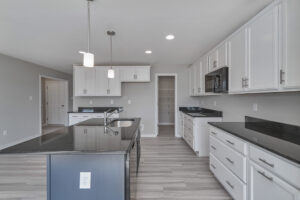 A modern kitchen with white cabinets, black countertops, stainless steel appliances, pendant lighting, and wooden flooring. A center island with a sink and an open pantry are also visible.