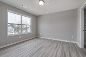 An empty room with light gray walls, wood flooring, a window with white trim, and a ceiling light fixture. A door on the right leads to a closet.