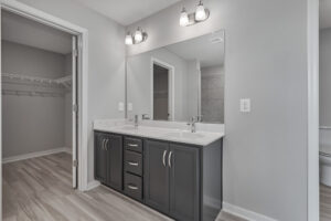 A bathroom with a dual-sink vanity, large mirror, gray cabinets, and light fixtures above. The room has a doorway leading to a walk-in closet with open shelving. The floor is light-colored tile.