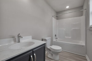 A modern bathroom with a dark vanity, white countertop, faucet, toilet, and a bathtub with a white surround and overhead light.