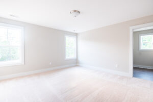 A bright, empty room with beige walls, carpeted floor, two windows, and a light fixture on the ceiling. A doorway leads to another room.