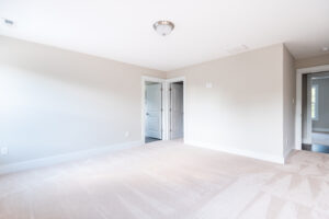 A bright, empty room with a beige carpet, white walls, a ceiling light fixture, and two closed white doors. A small section of a hallway is visible through an open door.