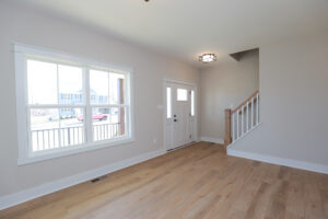 Bright room in Lot 1, featuring light wood floors, a large window, and a white front door beside the staircase.