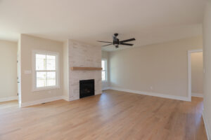 Empty living room with wooden floor, white brick fireplace, ceiling fan, and large windows in Poplar Villages Lot 1.