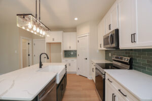 Modern kitchen in Poplar Village with white cabinets, green tile backsplash, and stainless steel appliances under a sleek light fixture.