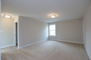 A spacious room in Poplar Village with beige carpet, beige walls, two ceiling lights, and a window.